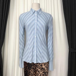 May include: A light blue long-sleeve button-down shirt featuring a repeating white floral pattern arranged in diagonal stripes. The shirt has a pointed collar, a central button placket with white buttons, and long sleeves with wide cuffs.