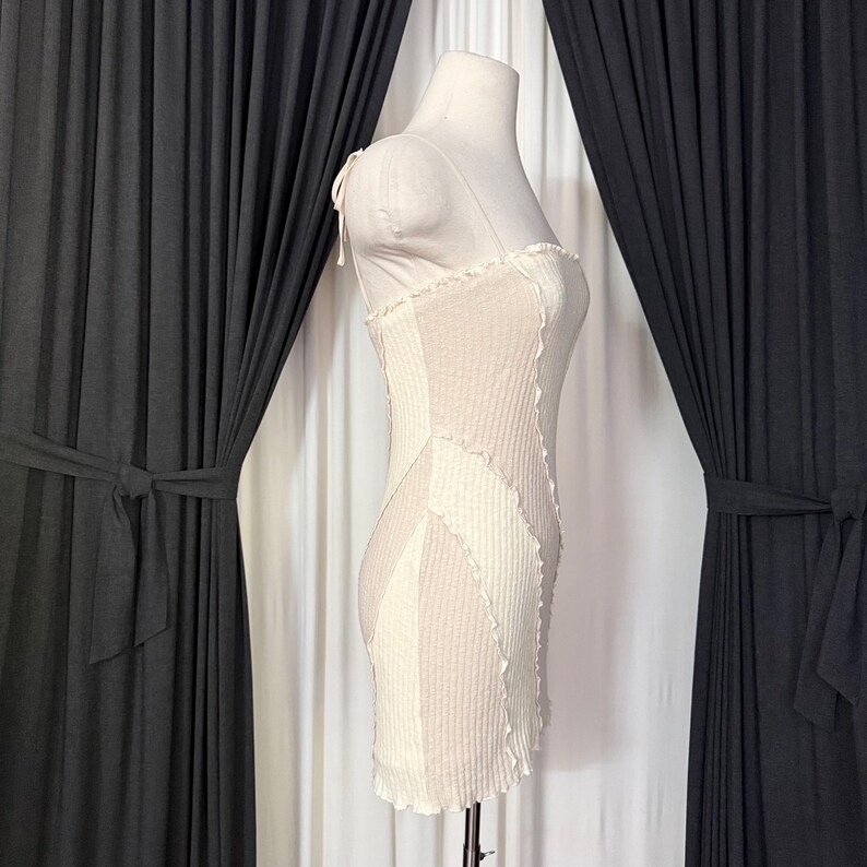 May include: A cream-colored, ribbed mini dress with a patchwork design. The dress features thin straps and a ruffled hem. The dress is displayed on a mannequin. The dress is approximately 32 inches long.