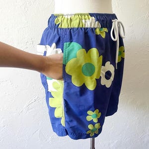 K&ouml;nnte beinhalten: Blaue Shorts mit einem Blumenmuster in Wei&szlig;, Gr&uuml;n und Gelb. Die Shorts haben einen Kordelzug in der Taille und Taschen. Die Shorts werden auf einer Schaufensterpuppe pr&auml;sentiert.