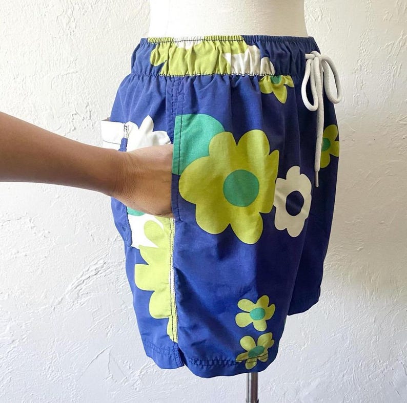 K&ouml;nnte beinhalten: Blaue Shorts mit einem Blumenmuster in Wei&szlig;, Gr&uuml;n und Gelb. Die Shorts haben einen Kordelzug in der Taille und Taschen. Die Shorts werden auf einer Schaufensterpuppe pr&auml;sentiert.