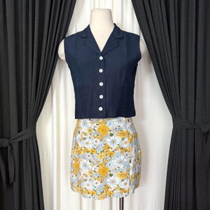 May include: A navy blue sleeveless top with white buttons paired with a floral mini skirt. The skirt features a pattern of yellow and white flowers on a light blue background. The top has a collared neckline.
