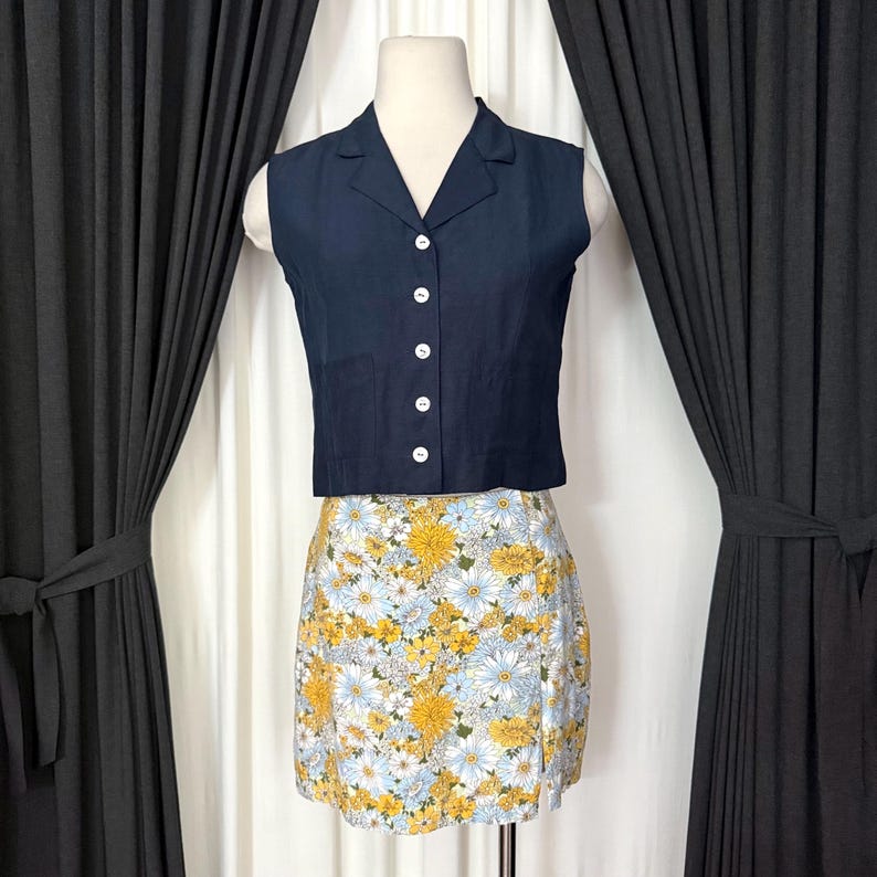 May include: A navy blue sleeveless top with white buttons paired with a floral mini skirt. The skirt features a pattern of yellow and white flowers on a light blue background. The top has a collared neckline.