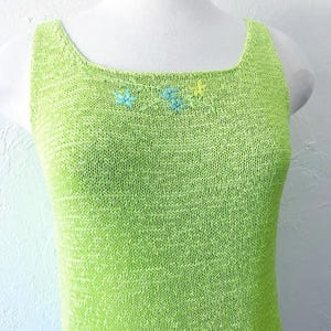 K&ouml;nnte beinhalten: Ein limettengr&uuml;nes Strick-Tanktop mit quadratischem Ausschnitt. Das Oberteil hat eine kleine Blumenstickerei in Blau- und Gelbt&ouml;nen am Ausschnitt. Das &auml;rmellose Oberteil wird auf einer wei&szlig;en Schaufenquin pr&auml;sentiert.