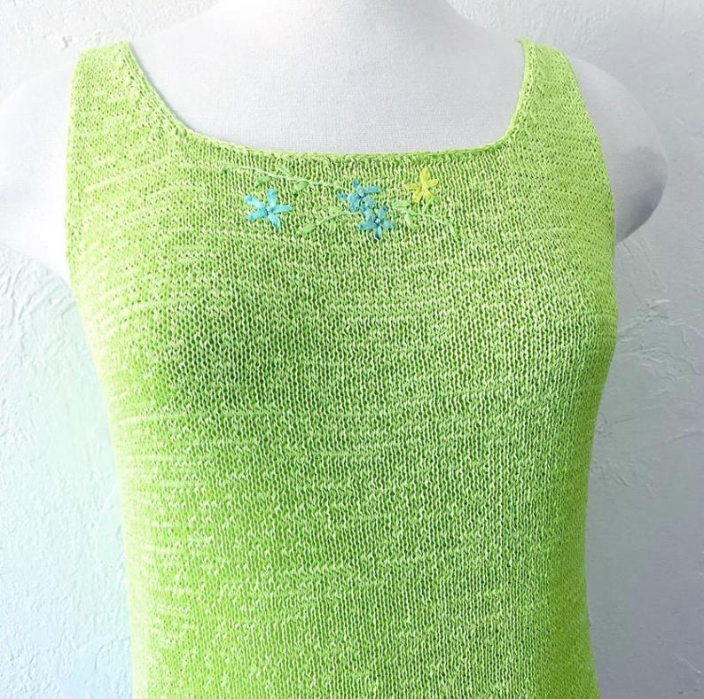 K&ouml;nnte beinhalten: Ein limettengr&uuml;nes Strick-Tanktop mit quadratischem Ausschnitt. Das Oberteil hat eine kleine Blumenstickerei in Blau- und Gelbt&ouml;nen am Ausschnitt. Das &auml;rmellose Oberteil wird auf einer wei&szlig;en Schaufenquin pr&auml;sentiert.