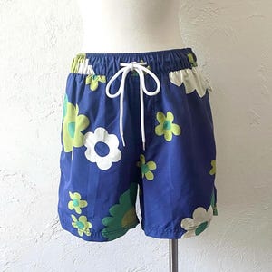 K&ouml;nnte beinhalten: Blaue Shorts mit wei&szlig;em Kordelzug und einem Blumenmuster in Wei&szlig;, Gr&uuml;n und Gelb. Die Shorts haben einen elastischen Bund und sind aus leichtem Material. Das Blumenmuster ist ein sich wiederholendes Muster aus gro&szlig;en und kleinen Blumen.