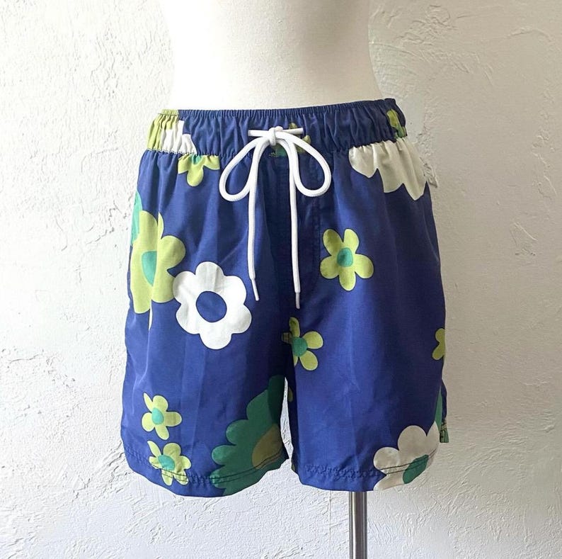 K&ouml;nnte beinhalten: Blaue Shorts mit wei&szlig;em Kordelzug und einem Blumenmuster in Wei&szlig;, Gr&uuml;n und Gelb. Die Shorts haben einen elastischen Bund und sind aus leichtem Material. Das Blumenmuster ist ein sich wiederholendes Muster aus gro&szlig;en und kleinen Blumen.