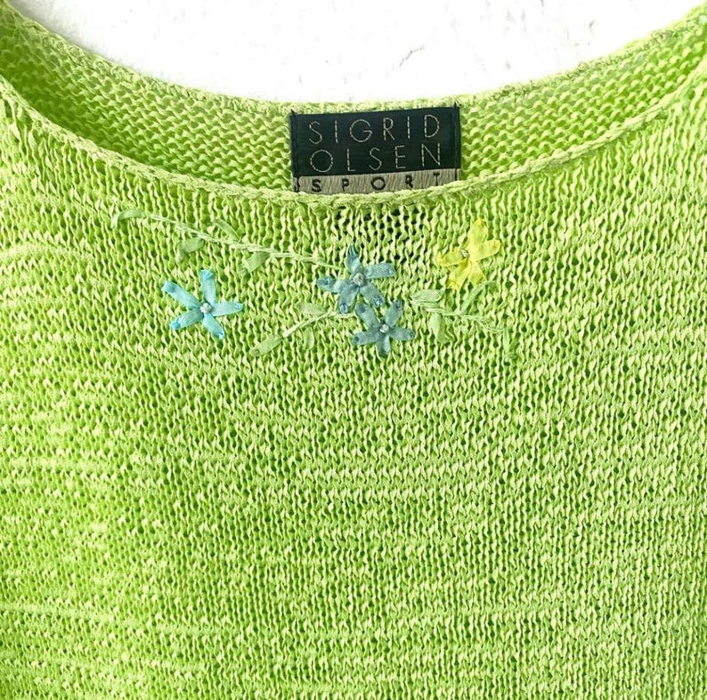 K&ouml;nnte beinhalten: Ein limettengr&uuml;nes Strickoberteil mit Rundhalsausschnitt. Das Oberteil hat gestickte Blumenakzente in Blau und Gelb. Das Etikett lautet "SIGRID OLSEN SPORT".