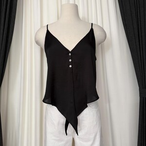 May include: Black camisole top with a V-neckline and spaghetti straps. The top features three white buttons down the center and a unique asymmetrical hemline with a tie detail. The top is displayed on a mannequin.
