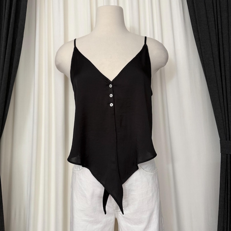 May include: Black camisole top with a V-neckline and spaghetti straps. The top features three white buttons down the center and a unique asymmetrical hemline with a tie detail. The top is displayed on a mannequin.