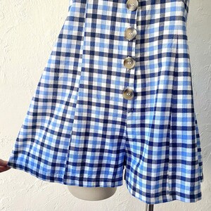 Puede incluir: Un mono de bot&oacute;n de gingham azul y blanco con falda corta y pantalones cortos. El mono tiene un cierre de bot&oacute;n en la parte delantera.