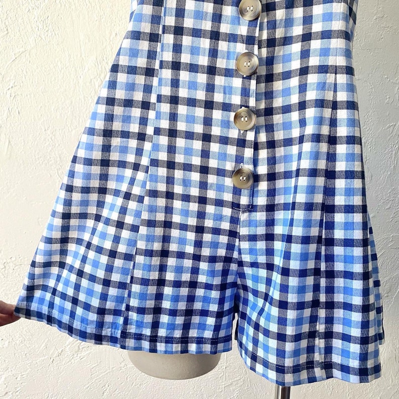 Puede incluir: Un mono de bot&oacute;n de gingham azul y blanco con falda corta y pantalones cortos. El mono tiene un cierre de bot&oacute;n en la parte delantera.