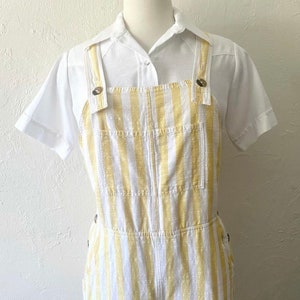 May include: White and yellow striped overalls with a white collared shirt. The overalls have a button closure and two pockets on the front.