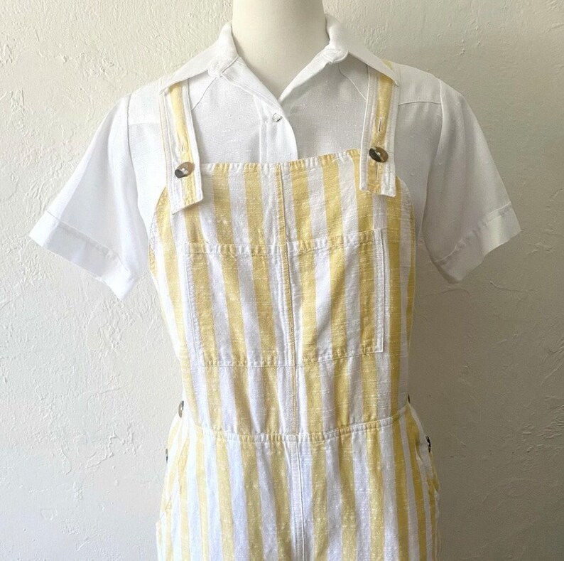May include: White and yellow striped overalls with a white collared shirt. The overalls have a button closure and two pockets on the front.