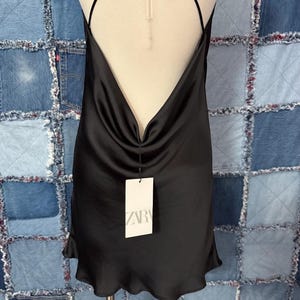 May include: A black satin slip dress with a cowl neckline and spaghetti straps. The dress has a low back and a slightly ruffled hem. A white tag with the brand name "ZARA" is attached.