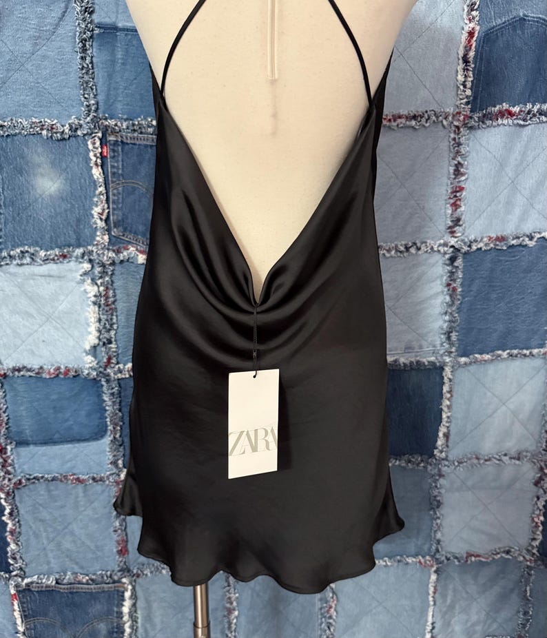 May include: A black satin slip dress with a cowl neckline and spaghetti straps. The dress has a low back and a slightly ruffled hem. A white tag with the brand name "ZARA" is attached.