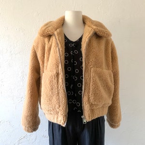 K&ouml;nnte beinhalten: Eine braune Kunstfelljacke mit Rei&szlig;verschluss und zwei Taschen. Die Jacke hat eine verk&uuml;rzte Passform und einen Kragen.