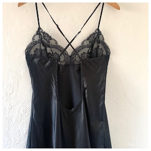 May include: Black satin slip dress with delicate spaghetti straps and a lace neckline. The back of the dress features a low scoop design. The dress is hanging on a wooden hanger against a white wall.