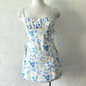 May include: White mini dress with a blue and green floral print. The dress has thin straps and a fitted bodice. The skirt is slightly flared.