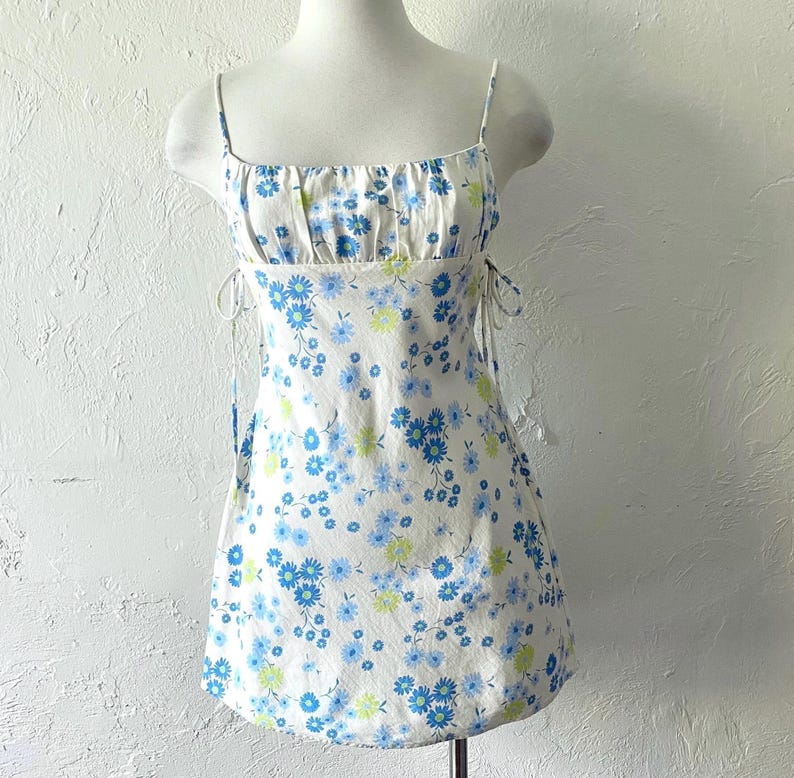 May include: White mini dress with a blue and green floral print. The dress has thin straps and a fitted bodice. The skirt is slightly flared.