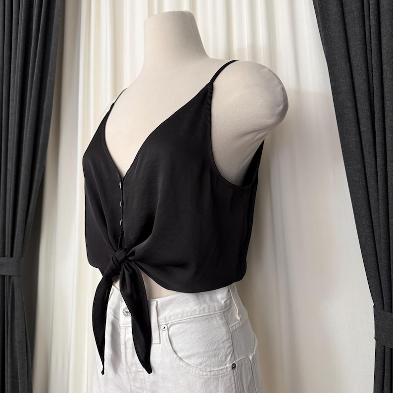 May include: A black camisole top with a V-neckline and spaghetti straps. The top features a tie detail at the waist and is displayed on a mannequin. The fabric appears to be a smooth, silky material.