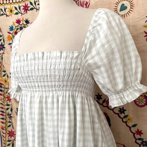 May include: A white and light green gingham dress with a square neckline and short puff sleeves. The dress has a smocked bodice and a flowing skirt.
