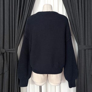May include: A navy blue knit sweater with a crew neck and long sleeves. The sweater has a textured knit pattern and is displayed on a mannequin. The sleeves are slightly puffed at the shoulders.