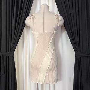 May include: A beige and cream-colored mini dress on a mannequin. The dress features a ribbed texture and a diagonal design. It has thin straps tied into bows at the shoulders and a ruffled hemline. The dress is displayed against a backdrop of dark curtains.