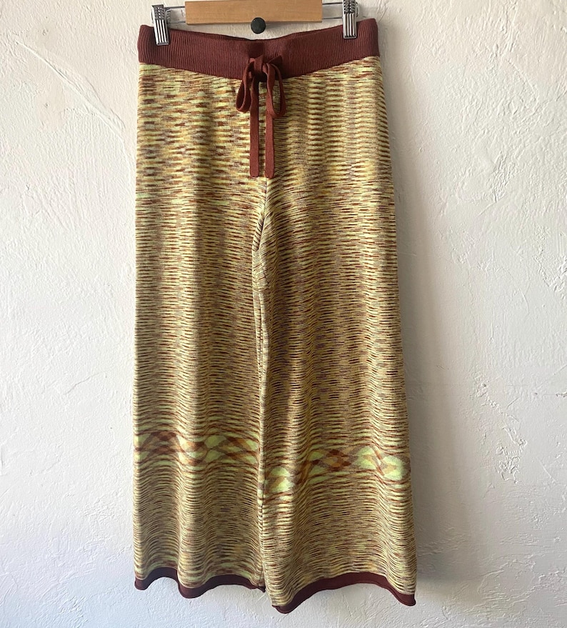 May include: Wide-leg knit pants with a brown waistband and a tie at the waist. The pants have a yellow and brown striped pattern with a geometric design at the bottom.