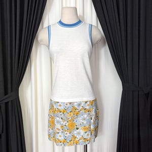 May include: A white sleeveless top with blue trim and a floral print mini skirt. The skirt features a pattern of yellow and blue flowers on a light blue background. The outfit is displayed on a mannequin.