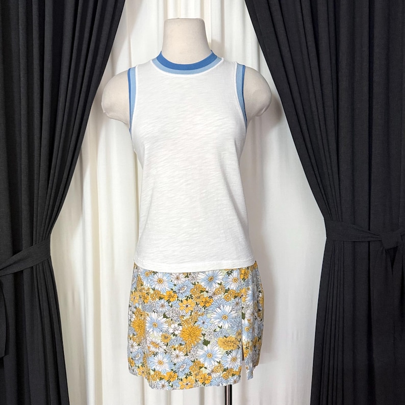 May include: A white sleeveless top with blue trim and a floral print mini skirt. The skirt features a pattern of yellow and blue flowers on a light blue background. The outfit is displayed on a mannequin.