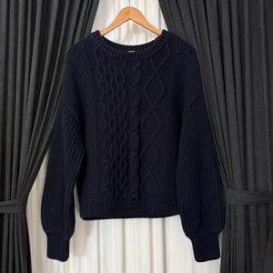 May include: A navy blue cable-knit sweater with a crew neck and long sleeves. The sweater features a textured cable-knit design down the center and on the sleeves. The sweater is hanging on a wooden hanger against a dark background.