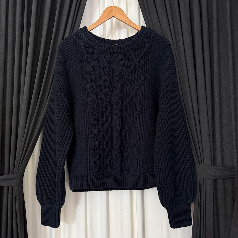 May include: A navy blue cable-knit sweater with a crew neck and long sleeves. The sweater features a textured cable-knit design down the center and on the sleeves. The sweater is hanging on a wooden hanger against a dark background.