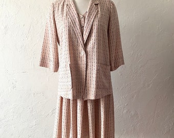 May include: A two-piece skirt suit in a pale pink with a delicate white and blue patterned design. The jacket has a single button closure and a pocket on the left side. The skirt is pleated and falls to just above the knee.