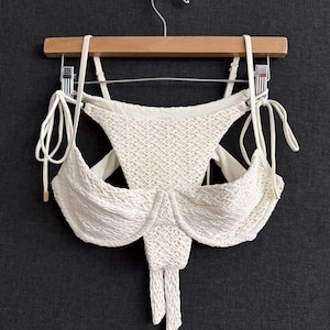 Peut inclure: Un ensemble de bikini blanc avec un motif tissé texturé. Le haut présente un soutien-gorge à armatures et des bretelles réglables. Le bas est taille haute avec un motif tissé. L'ensemble est présenté sur un cintre en bois.