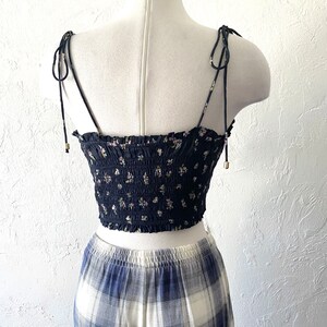 May include: A navy blue crop top with a floral pattern, featuring adjustable tie straps. The top is paired with a blue and white plaid skirt, creating a coordinated outfit. The top has a smocked bodice and delicate floral print.