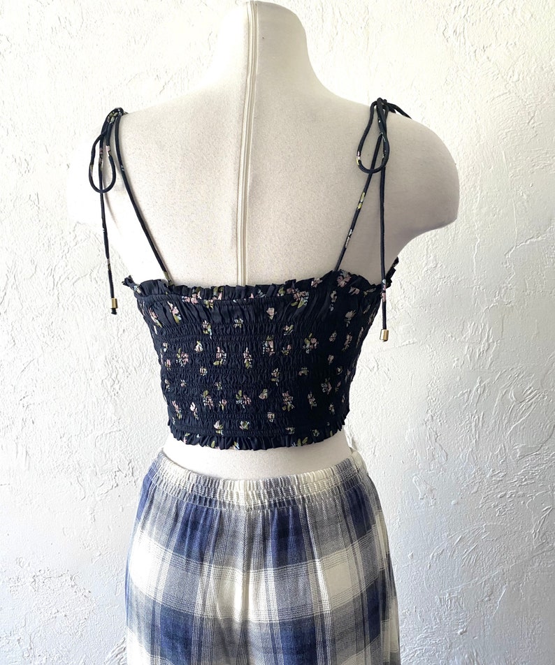 May include: A navy blue crop top with a floral pattern, featuring adjustable tie straps. The top is paired with a blue and white plaid skirt, creating a coordinated outfit. The top has a smocked bodice and delicate floral print.