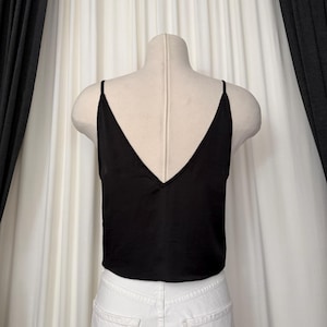 May include: A black camisole top with a deep V-neck and spaghetti straps. The top is displayed on a mannequin, paired with white denim jeans. The fabric appears to be a smooth, possibly silky material. The background features white curtains and dark gray drapes.