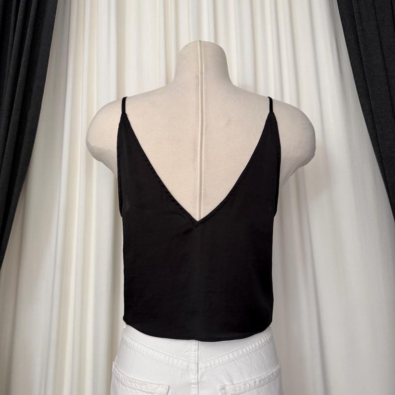 May include: A black camisole top with a deep V-neck and spaghetti straps. The top is displayed on a mannequin, paired with white denim jeans. The fabric appears to be a smooth, possibly silky material. The background features white curtains and dark gray drapes.