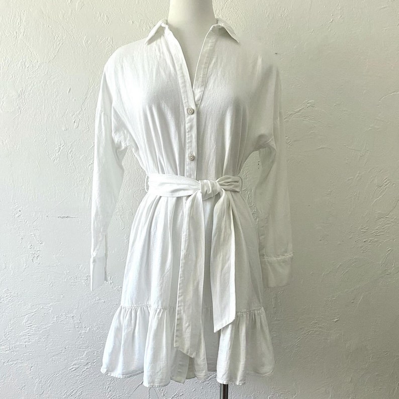 Puede incluir: Un vestido blanco de lino con botones, mangas largas y cintur&oacute;n de lazo. El vestido tiene un dobladillo con volantes.