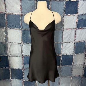 May include: A black satin slip dress with a cowl neckline and spaghetti straps. The dress is displayed on a mannequin against a patchwork denim background. The dress has a simple, elegant design.