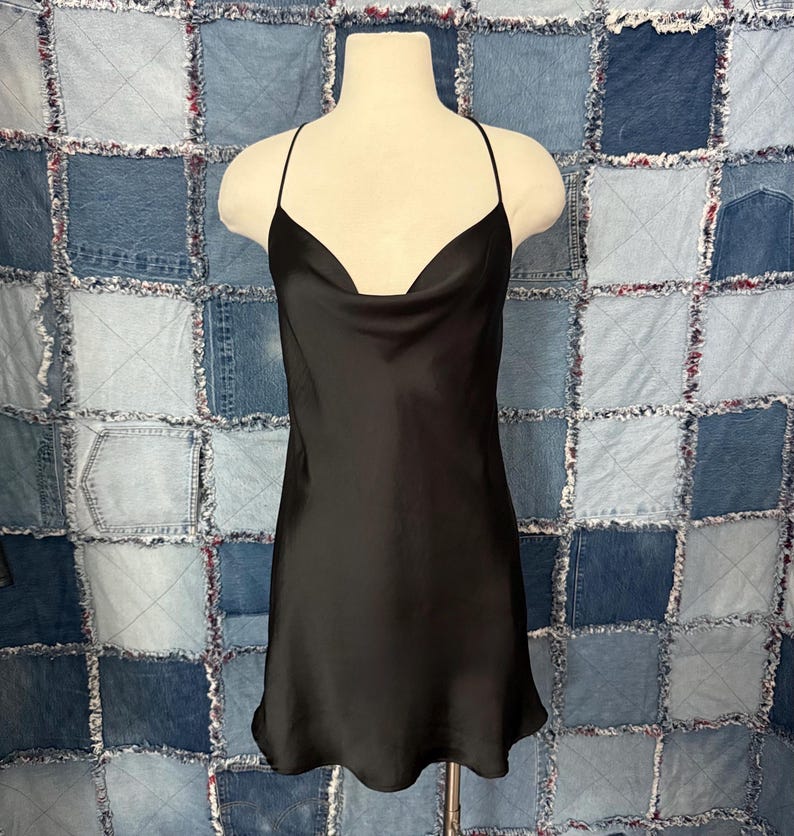 May include: A black satin slip dress with a cowl neckline and spaghetti straps. The dress is displayed on a mannequin against a patchwork denim background. The dress has a simple, elegant design.