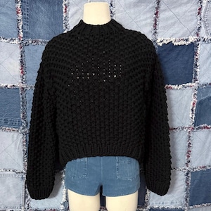 May include: A black, chunky knit sweater with a mock neck. The sweater has long sleeves and a cropped fit. The garment is displayed on a mannequin, paired with denim shorts. The background is a patchwork of denim fabric.