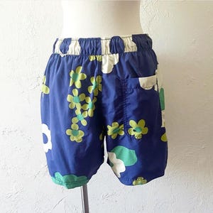 K&ouml;nnte beinhalten: Blaue Shorts mit einem Blumenmuster aus wei&szlig;en, gr&uuml;nen und gelben Bl&uuml;ten. Sie haben einen elastischen Bund und eine einzelne Tasche. Der Stoff scheint leicht zu sein, ideal f&uuml;r warmes Wetter. Die Shorts sind etwa 30 cm lang.
