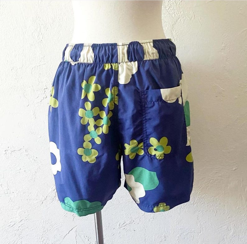 K&ouml;nnte beinhalten: Blaue Shorts mit einem Blumenmuster aus wei&szlig;en, gr&uuml;nen und gelben Bl&uuml;ten. Sie haben einen elastischen Bund und eine einzelne Tasche. Der Stoff scheint leicht zu sein, ideal f&uuml;r warmes Wetter. Die Shorts sind etwa 30 cm lang.
