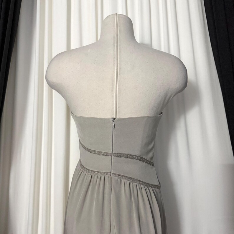 May include: A strapless light gray dress displayed on a mannequin. The garment features a fitted bodice with a central vertical zipper closure and two horizontal bands of intricate lace trim detailing that wrap around the waist area.