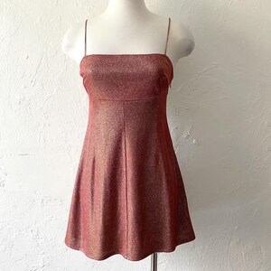 Puede incluir: Un minivestido rojo-marr&oacute;n brillante con finas correas de espagueti. El vestido tiene un escote cuadrado y un corpi&ntilde;o ajustado que se ensancha en una falda en forma de A. La tela tiene una textura brillante.