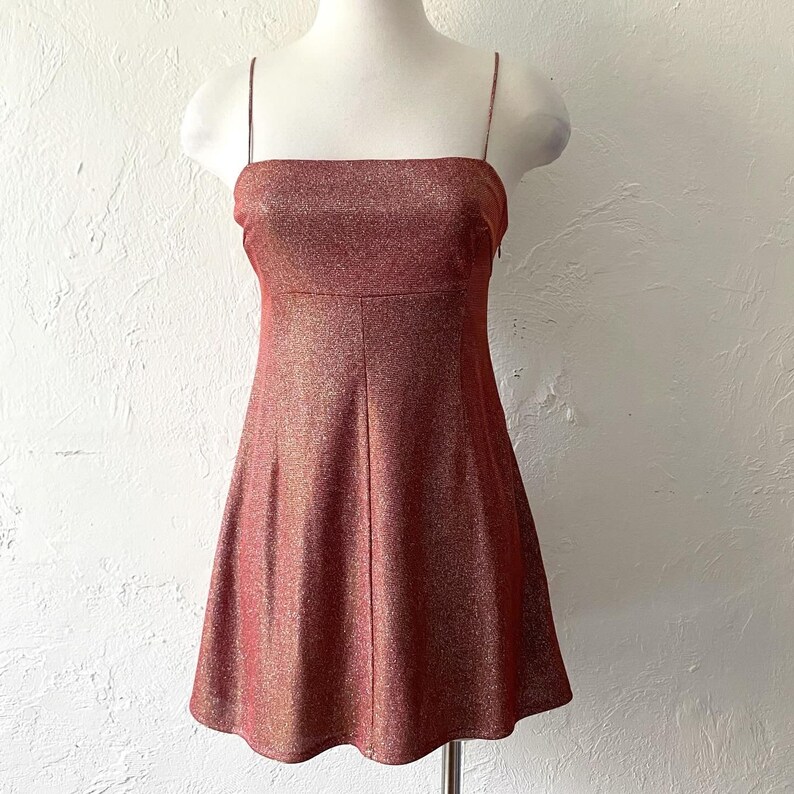 Puede incluir: Un minivestido rojo-marr&oacute;n brillante con finas correas de espagueti. El vestido tiene un escote cuadrado y un corpi&ntilde;o ajustado que se ensancha en una falda en forma de A. La tela tiene una textura brillante.