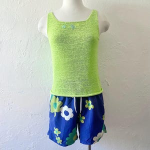 K&ouml;nnte beinhalten: Ein limettengr&uuml;nes Tanktop mit quadratischem Ausschnitt und kleinen floralen Verzierungen, kombiniert mit blauen Shorts mit Blumenmuster. Die Shorts haben einen wei&szlig;en Kordelzug und ein gr&uuml;n-wei&szlig;es Blumenmuster.