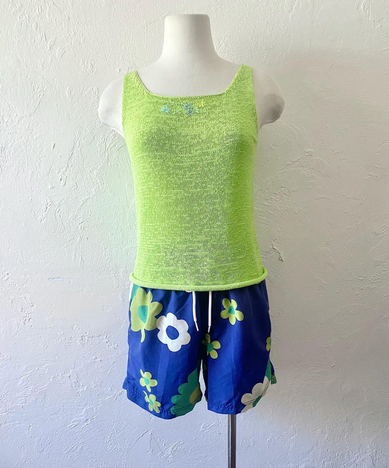 K&ouml;nnte beinhalten: Ein limettengr&uuml;nes Tanktop mit quadratischem Ausschnitt und kleinen floralen Verzierungen, kombiniert mit blauen Shorts mit Blumenmuster. Die Shorts haben einen wei&szlig;en Kordelzug und ein gr&uuml;n-wei&szlig;es Blumenmuster.