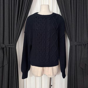 May include: A navy blue cable-knit sweater with a crew neck. The sweater features a textured pattern on the front and sleeves. The sleeves are long and the hemline is ribbed. The sweater is displayed on a mannequin.